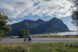 A serene landscape featuring a majestic mountain range under a partly cloudy sky, with a calm body of water at the base. In the foreground, a touring bicycle loaded with gear is parked by the roadside, surrounded by sparse greenery.