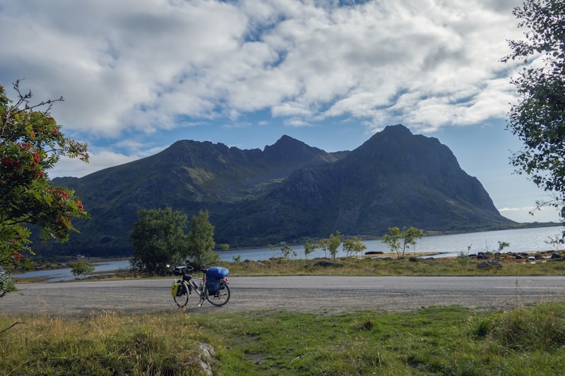 A serene landscape featuring a majestic mountain range under a partly cloudy sky, with a calm body of water at the base. In the foreground, a touring bicycle loaded with gear is parked by the roadside, surrounded by sparse greenery.