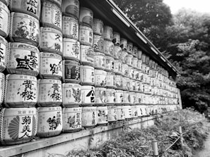 Rows of traditional sake barrels stacked along a wall, displaying intricate calligraphy and designs. The barrels are arranged artistically, conveying cultural significance. The background includes lush green foliage, adding a natural element.