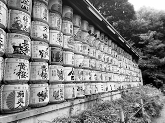 Rows of traditional sake barrels stacked along a wall, displaying intricate calligraphy and designs. The barrels are arranged artistically, conveying cultural significance. The background includes lush green foliage, adding a natural element.