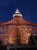 An impressive view of a lattice tower structure, likely a broadcasting tower. The complex network of red-orange steel beams is illuminated against the dark evening sky. The top of the tower is adorned with lights, creating a contrast with the deep blue background.