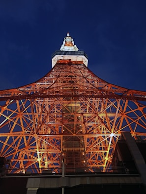 An impressive view of a lattice tower structure, likely a broadcasting tower. The complex network of red-orange steel beams is illuminated against the dark evening sky. The top of the tower is adorned with lights, creating a contrast with the deep blue background.