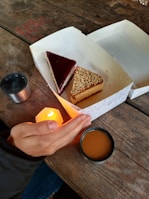 Two slices of cake are placed inside a white box on a rustic wooden table. One slice has a dark red glossy topping, while the other is covered with chopped nuts. A hand is positioned near a small candle, bringing a sense of warmth to the scene. A metal cup filled with a hot beverage is also on the table.