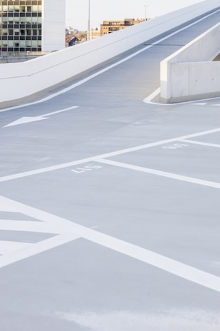 A modern, empty parking lot with marked spaces and a smooth concrete surface. White arrows and lines indicate directions and parking boundaries. The area is well-lit with clean architectural lines, and in the background, a multi-story building and urban elements are visible.
