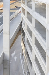 A tall, multi-story parking structure with metal mesh panels on one side, adjacent to a highway with several cars in motion. The scene includes concrete walls and ramps, with directional arrows painted on the ground.