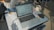 A laptop is open on a desk, displaying a website or application interface. Surrounding the laptop are various items including a mug, a face mask, a snack pack, and some stationery. A person is seated at the desk, engaged with a device, possibly a phone, though their face isn't visible. The setting appears to be a classroom or study space due to the presence of chairs and desks.