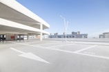 A spacious and empty parking lot with white lane markings and arrows on a concrete surface. The area is open-air with a modern building featuring a grid-like facade in the background. Two sleek, minimalist light poles are positioned in the center of the scene, and the sky is clear and blue.