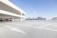 A spacious and empty parking lot with white lane markings and arrows on a concrete surface. The area is open-air with a modern building featuring a grid-like facade in the background. Two sleek, minimalist light poles are positioned in the center of the scene, and the sky is clear and blue.