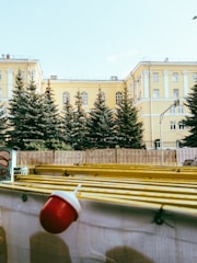 A large yellow building with multiple windows and classical architecture stands behind a row of evergreen trees. In the foreground, there is a construction barrier decorated with graffiti and topped with a yellow structure and a red and white light fixture.