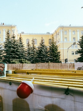A large yellow building with multiple windows and classical architecture stands behind a row of evergreen trees. In the foreground, there is a construction barrier decorated with graffiti and topped with a yellow structure and a red and white light fixture.