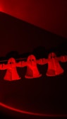 Three paper ghost cutouts with arms extended, illuminated in red light, are suspended in a dark setting. Each ghost has a simple design with large black eyes and a flowing robe-like shape.