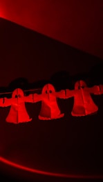 Three paper ghost cutouts with arms extended, illuminated in red light, are suspended in a dark setting. Each ghost has a simple design with large black eyes and a flowing robe-like shape.