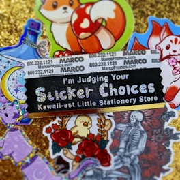 A collection of colorful and whimsical stickers displayed on a glittery gold background. The central black sticker with glittery text reads 'I'm Judging Your Sticker Choices' followed by 'Kawaii-est Little Stationery Store.' Surrounding this sticker are various images, including a fox with a red and white mushroom, a bottle with ribbons, a crescent moon with stars, a purple cat, a bird amidst roses, and a monochrome skeletal figure with a bouquet of roses. Some promotional text and numbers appear on a white background towards the top.