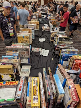 A crowded indoor event where people are browsing through a large selection of board games displayed on tables. The scene shows a diverse group of individuals engaging with the games, some are intently examining boxes, while others move through the aisles. The atmosphere is lively and social, with games organized in rows.