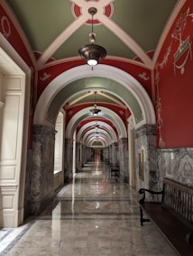A luxurious hallway featuring ornate arches and intricate designs on the ceiling and walls. The passage is made of polished marble with a glossy finish, enhanced by the symmetrical repetition of lamps hanging from the center of each arch. The colors and patterns create a sense of depth and elegance, with benches lining the walls.