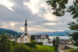 A picturesque scene of a serene village with a prominent church featuring a tall steeple and a spire. The area is lush with greenery and surrounded by a lake, hills, and distant mountains under a cloudy sky.