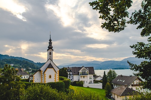 A picturesque scene of a serene village with a prominent church featuring a tall steeple and a spire. The area is lush with greenery and surrounded by a lake, hills, and distant mountains under a cloudy sky.