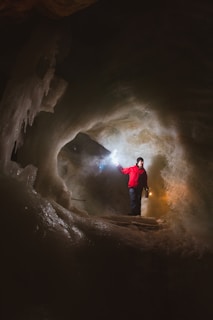A person clad in a bright red jacket stands inside an ice cave, holding a lantern in one hand and a flashlight in the other. The cave walls glisten with ice formations, illuminated by the lights. The atmosphere is mysterious and adventurous with shadows casting intricate patterns around the figure.