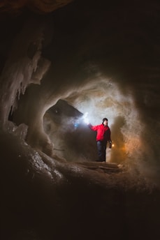A person clad in a bright red jacket stands inside an ice cave, holding a lantern in one hand and a flashlight in the other. The cave walls glisten with ice formations, illuminated by the lights. The atmosphere is mysterious and adventurous with shadows casting intricate patterns around the figure.
