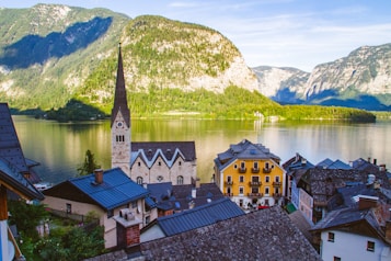 A picturesque village with traditional alpine architecture, nestled by a serene lake surrounded by majestic mountains. The focal point is a charming church with a tall spire, complemented by colorful buildings nearby. The tranquil water reflects the lush greenery and towering cliffs in the background.