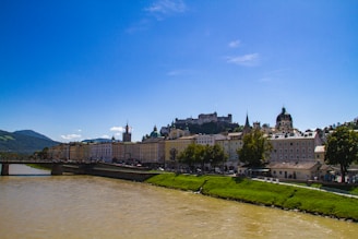 Buildings line the edge of a waterway with a historic fortress atop a hill in the background. The scene is set under a clear blue sky with mountains in the distance and greenery along the riverbank.