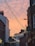 A narrow city street with buildings on either side, adorned with hanging string lights in the shape of stars. The sky features a warm, orange-pink hue, suggesting a sunset or sunrise. Signs for local businesses and colleges are visible on the buildings, including one for the Northern College of Costume.