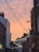A narrow city street with buildings on either side, adorned with hanging string lights in the shape of stars. The sky features a warm, orange-pink hue, suggesting a sunset or sunrise. Signs for local businesses and colleges are visible on the buildings, including one for the Northern College of Costume.
