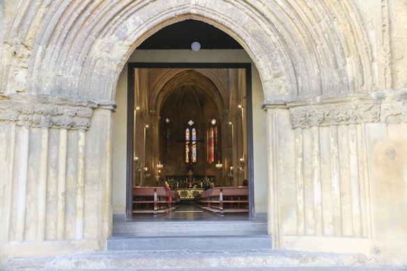 An arched stone entryway leads into the interior of a church featuring high ceilings, wooden pews, and colorful stained glass windows. The soft lighting adds a serene and spiritual atmosphere.