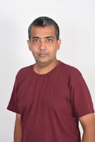 A person with short, dark hair is wearing a maroon t-shirt. They are standing against a plain white background, looking directly at the camera with a neutral expression.