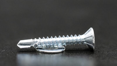 A close-up image focusing on a metallic screw resting on a dark surface. The screw has a flat head and a sharp, pointed tip with clear, visible threads that reflect light.