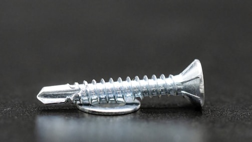 A close-up image focusing on a metallic screw resting on a dark surface. The screw has a flat head and a sharp, pointed tip with clear, visible threads that reflect light.