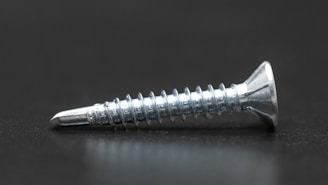 A single metallic screw with a shiny, reflective surface lies horizontally on a dark surface. It has a pointed tip and spiral threads along its body, with a flat, countersunk head.
