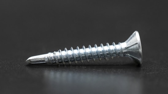 A single metallic screw with a shiny, reflective surface lies horizontally on a dark surface. It has a pointed tip and spiral threads along its body, with a flat, countersunk head.