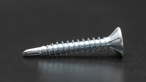 A single metallic screw with a shiny, reflective surface lies horizontally on a dark surface. It has a pointed tip and spiral threads along its body, with a flat, countersunk head.