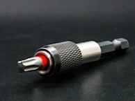 A close-up view of a metallic drill bit adapter featuring a textured, knurled grip and a visible red accent. The tool is placed on a dark surface, highlighting its polished metal components.