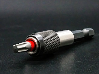 A close-up view of a metallic drill bit adapter featuring a textured, knurled grip and a visible red accent. The tool is placed on a dark surface, highlighting its polished metal components.