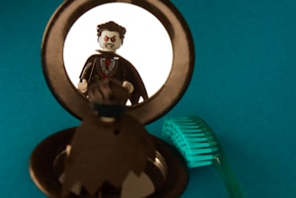 A LEGO figure dressed as a vampire looks at itself in a round mirror. The background is a solid teal color, and there is a green, translucent LEGO part on the right side of the image.