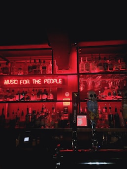 A dimly lit bar shelf is filled with various bottles of alcohol, illuminated by a strong red neon light. The neon sign reads 'MUSIC FOR THE PEOPLE' and casts a vibrant glow over the scene. On the counter, beer taps with decorative handles, including one with a skull motif, are visible.