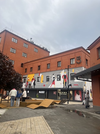 A modern urban scene featuring a brick building with a mural depicting stylized figures in vibrant colors. The scene shows a paved pedestrian area with wooden benches and a few people walking or standing. The sky above is overcast, giving a muted light to the setting.