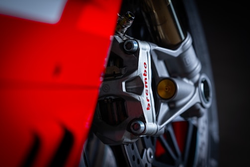 Close-up view of a motorcycle brake caliper with the brand name 'Brembo' in red lettering. The image features a metal structure with intricate details and a striking red background, highlighting the mechanical components.