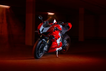 A sleek, red sport motorcycle is parked under dim lighting in an indoor space. The angular design of the bike is highlighted by the contrast between the red body and the black and white accents. The setting appears to be a parking garage with a warm, moody ambiance created by the surrounding orange and red tones.
