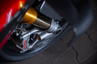 Close-up view of a high-performance motorcycle brake caliper. The Brembo branding is visible in red on the silver caliper, attached to a disc brake. The image also features parts of the suspension system with golden and silver components, against a dark wheel and tire background.