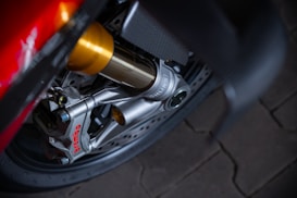 Close-up view of a high-performance motorcycle brake caliper. The Brembo branding is visible in red on the silver caliper, attached to a disc brake. The image also features parts of the suspension system with golden and silver components, against a dark wheel and tire background.