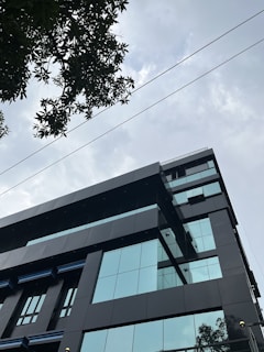 A modern office building with reflective glass windows set against a cloudy sky, partially obscured by tree branches in the foreground. The structure features sleek, dark facade panels and sharp geometric lines.