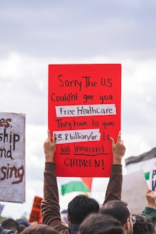 A group of people is participating in a protest. One prominent red sign held aloft reads: 'Sorry The U.S couldn't give you Free Healthcare. They have to give $3.8 billion/yr to kill innocent children!' Parts of other signs are visible in the background, and the sky appears overcast.
