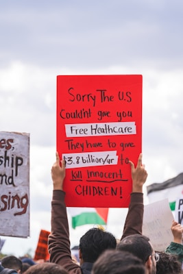 A group of people is participating in a protest. One prominent red sign held aloft reads: 'Sorry The U.S couldn't give you Free Healthcare. They have to give $3.8 billion/yr to kill innocent children!' Parts of other signs are visible in the background, and the sky appears overcast.