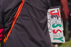 A person is holding a sign with bold, colorful text saying 'JEWS SAY' in red, black, and green. The person is wearing a dark shirt and has an orange strap over their shoulder.