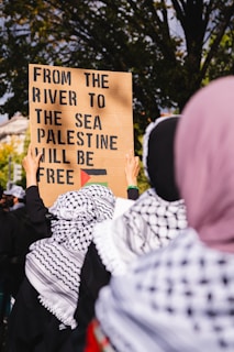 Several individuals wearing patterned scarves are gathered outdoors, holding a sign with bold text advocating for a cause. Trees and a building are visible in the background, suggesting a public demonstration or protest.