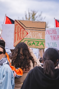 A group of individuals gathered at a protest or demonstration, holding signs with messages related to Palestine. One prominent sign is decorated with the words 'Free Palestine' and 'Gaza' in bold letters. Flags, possibly Palestinian, are visible in the background, and the crowd seems engaged in a social or political cause.