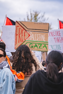A group of individuals gathered at a protest or demonstration, holding signs with messages related to Palestine. One prominent sign is decorated with the words 'Free Palestine' and 'Gaza' in bold letters. Flags, possibly Palestinian, are visible in the background, and the crowd seems engaged in a social or political cause.